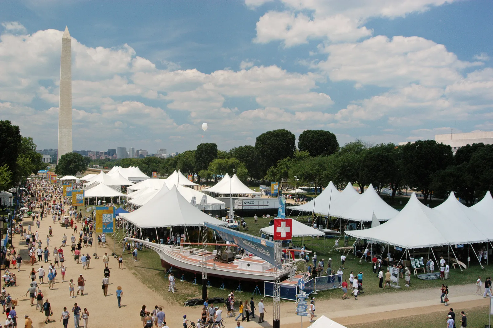 White peaked festival tents and crowds spread across the National Mall during the Smithsonian Folklife Festival, the Washington Monument rising behind, in Washington, D.C.