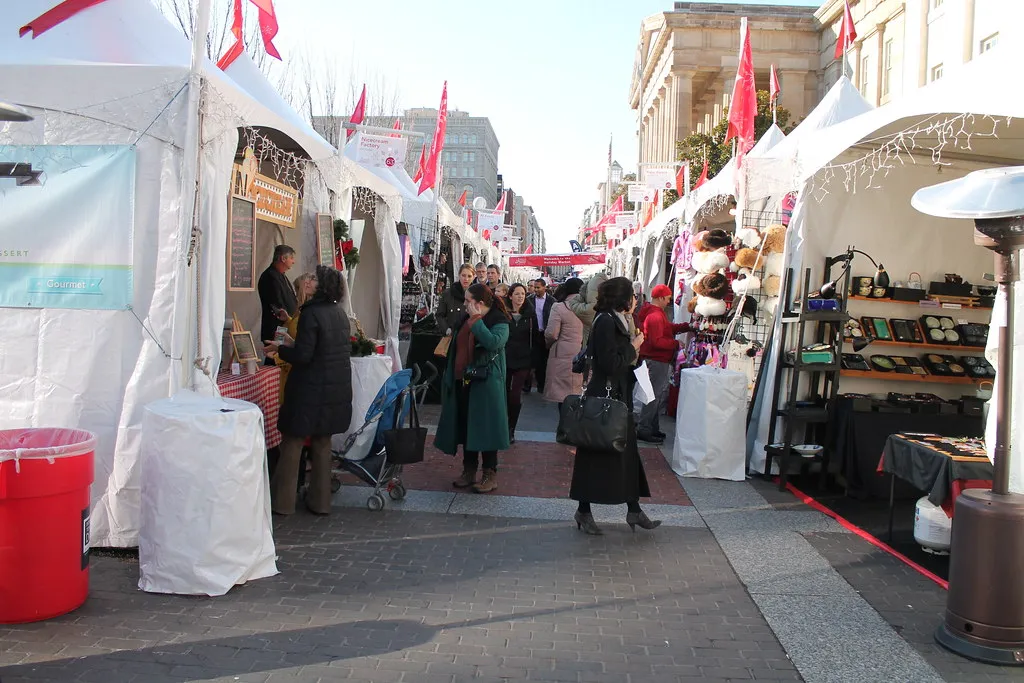 Rows of white-tented stalls with red flags down F Street NW at the Downtown Holiday Market, shoppers browsing in winter coats, in Washington, D.C.